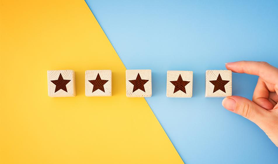 Five wooden blocks are shown against a half-yellow, half-pastel blue background. Stars are detailed on the blocks, and a hand places the 5th star block in place. 