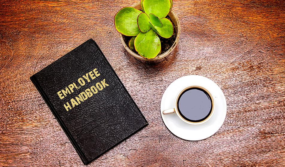 An employee handbook is on a dark brown, wooden table with black coffee in a cup and a plant next to it.