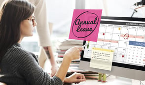 A woman is looking at an annual leave calendar on her screen, pointing at it with a pen. She seems to be responsible for staff holiday scheduling.