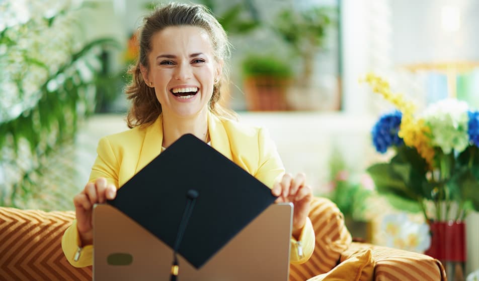 A smiling young woman is shown, balancing her graduation mortarboard on the top of her laptop.