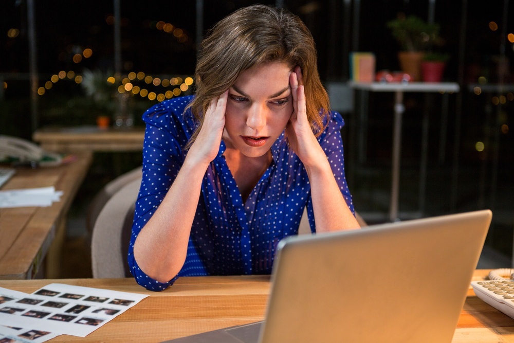 A stressed business woman is wearing a bright blue blouse and is sitting at a desk with her hands on her head. The woman is looking at her laptop in front of her and appears stressed. 