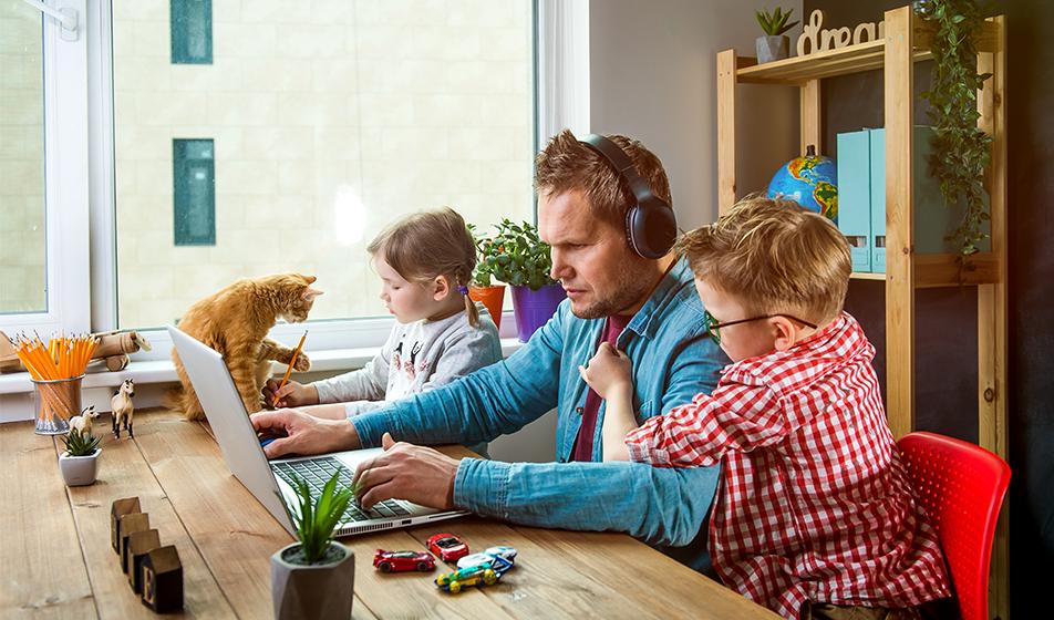 A man works from home on a laptop. Two children are either side of him, one is clambering on him. A cat is also on the table. 
