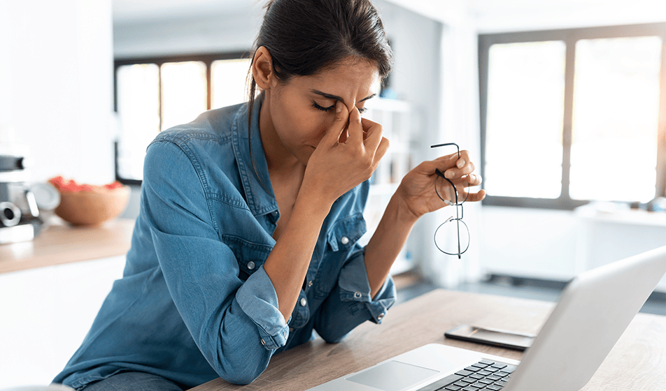 A woman sits at her kitchen counter with her laptop in front of her, working from home. She has taken her glasses off and holds them in one hand. Her eyes are closed and she's pinching the top of her nose near her forehead with her other hand. She looks stressed. 